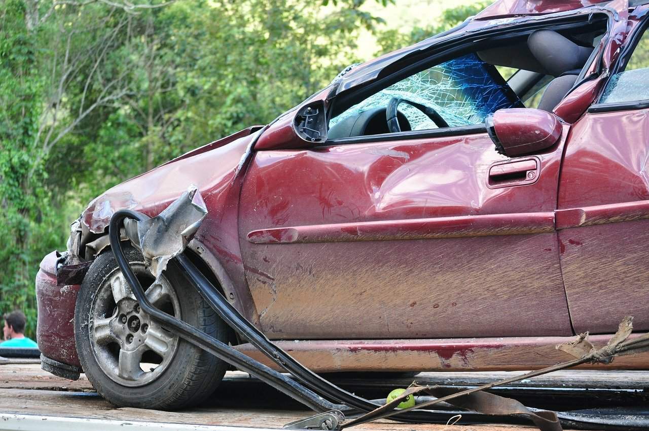 A dark red car with dents, missing parts, and broken windows after an accident.