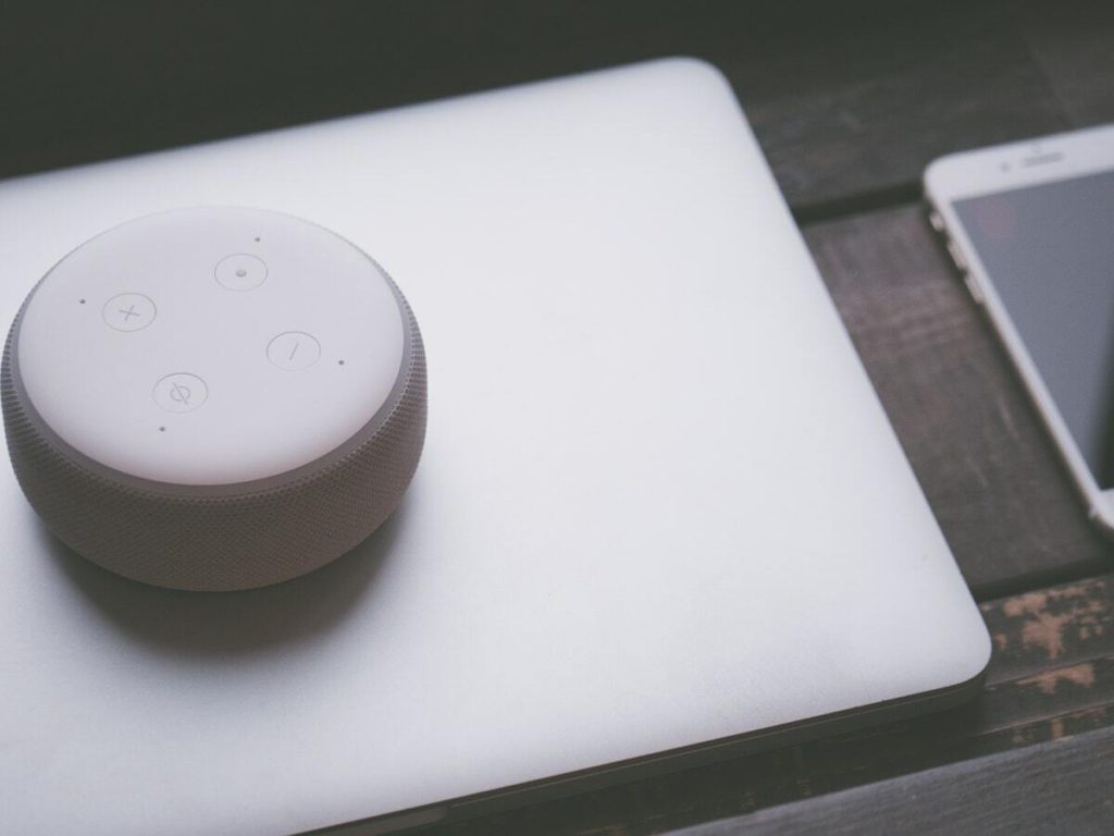 A white Amazon Echo Dot sits on top of a silver laptop in a rural setting.