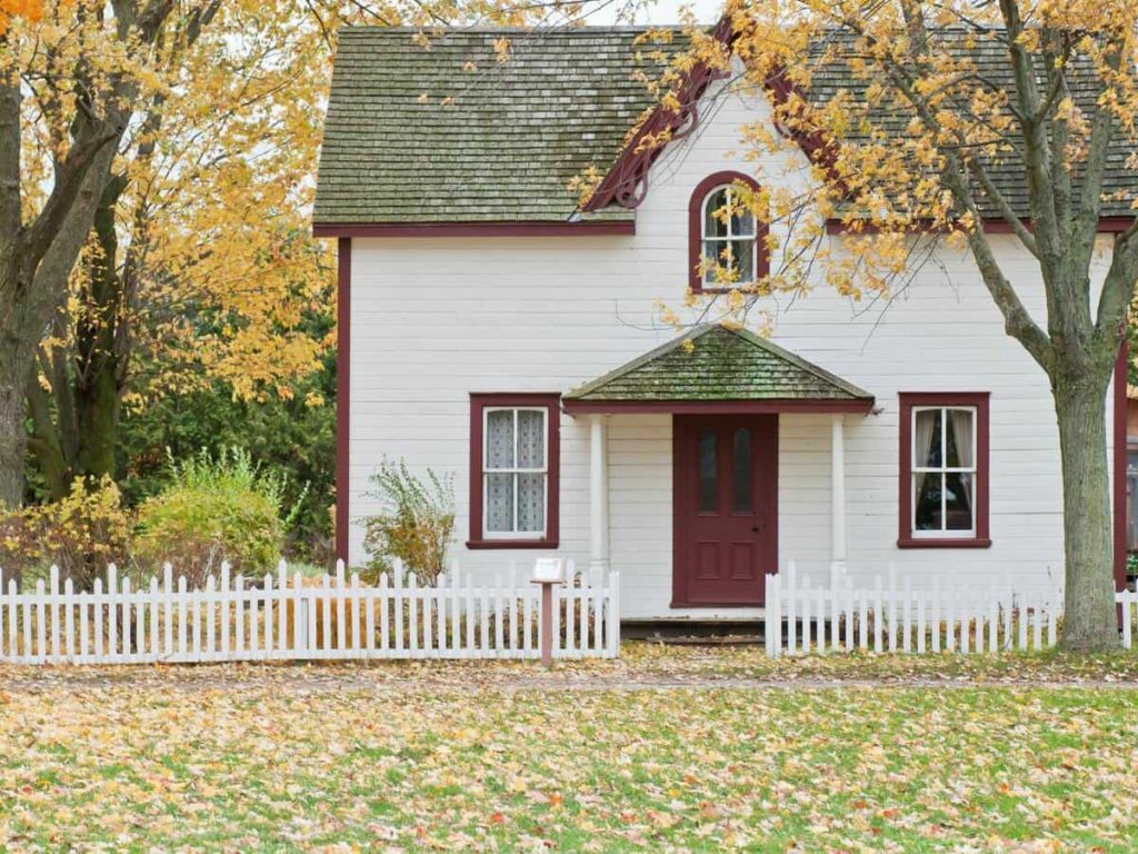 A small, two-story home with white paneling is next to trees with orange leaves in the fall.