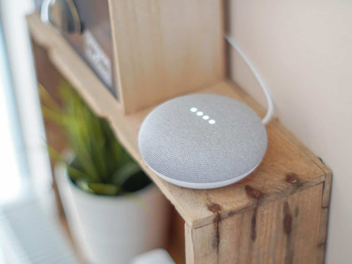 A grey google home mini sitting on top of a brown wooden bookcase.