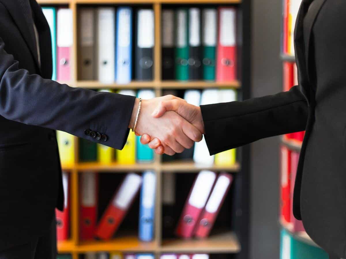 Two people in professional clothing are shaking hands in a working environment.