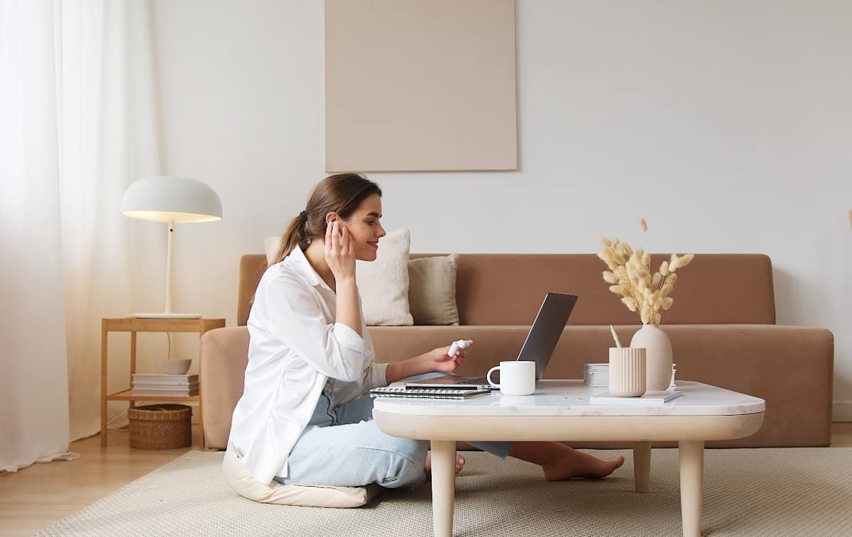 A young woman sitting on the floor of her living room as she puts on her cordless headphones and works from her laptop.