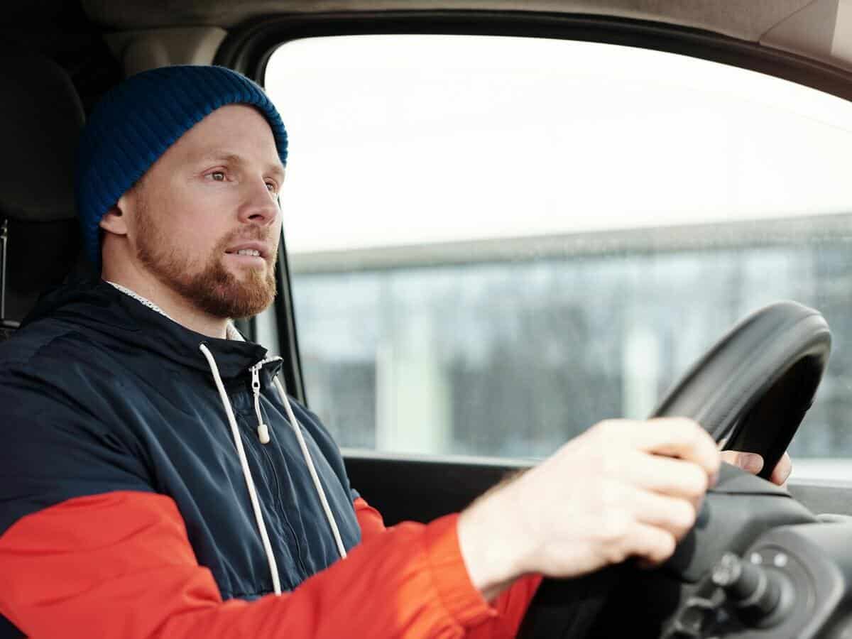 A man dressed in winter clothes has his hands on a black steering wheel as he drives a vehicle.