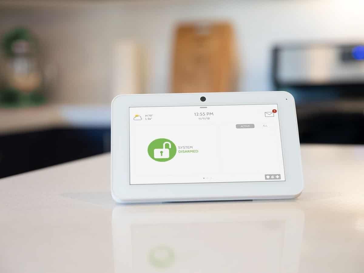 A white Qolsys IQ Panel 2 sits on top of a white granite countertop inside of a kitchen.