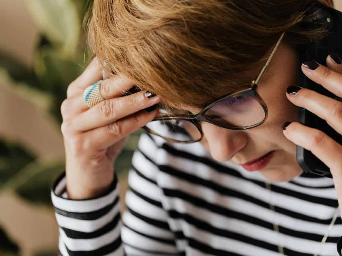 A woman holding the top of her head while looking down and listening on the phone.