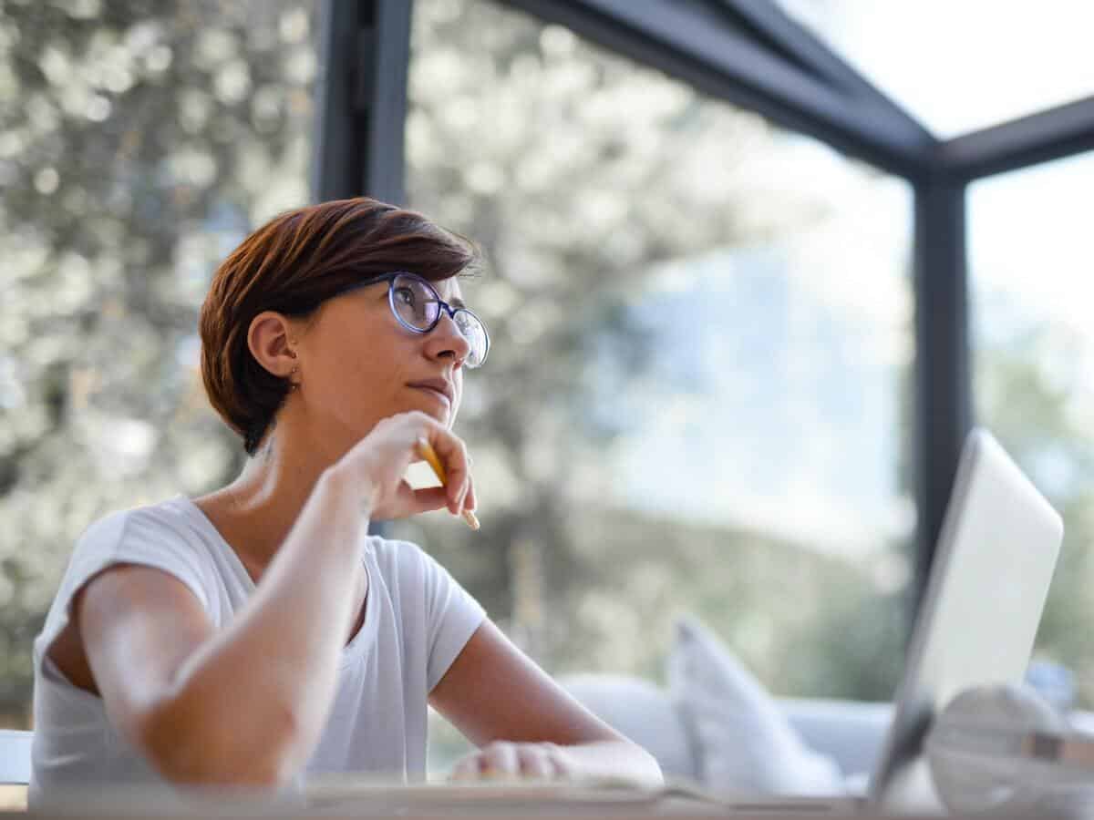 A pondering woman wearing glasses is sitting in front of her laptop.