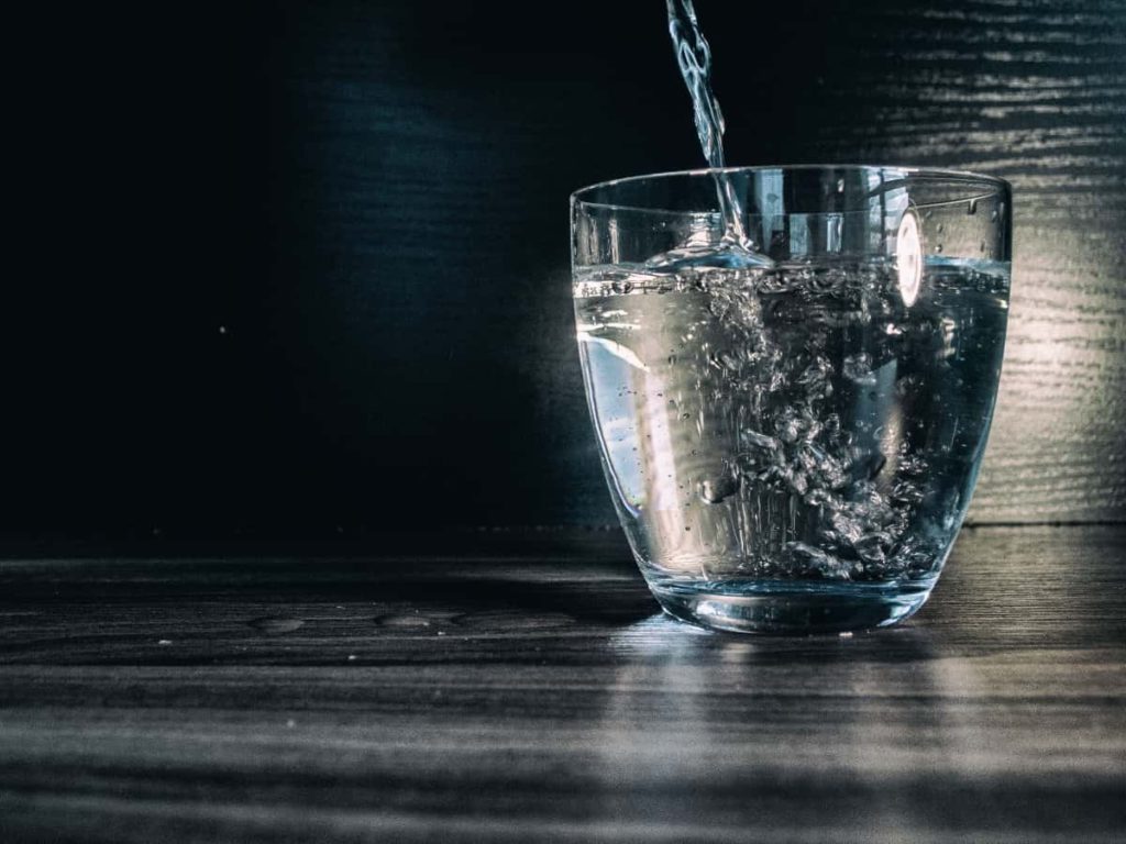 Water pouring into a modern, glass cup sitting on a dark wooden surface.