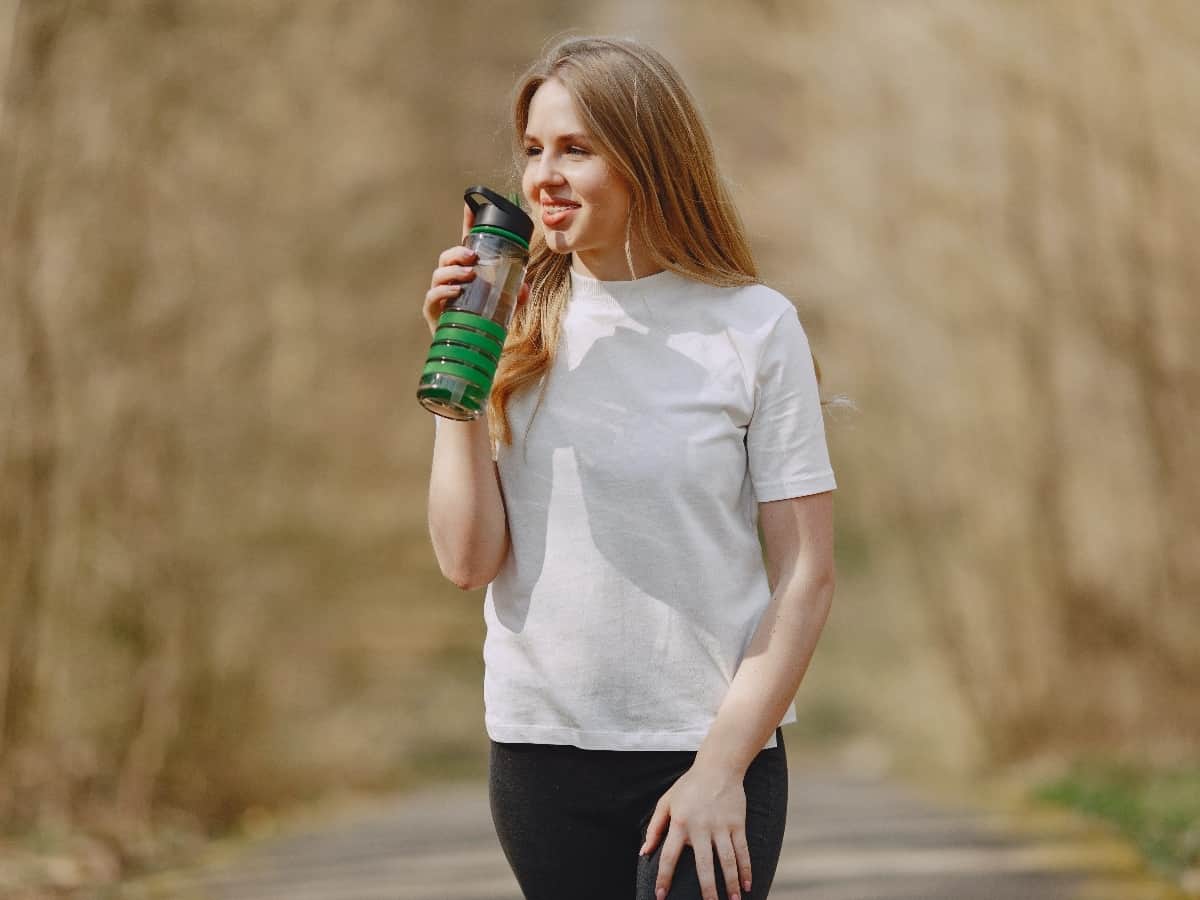 woman with light brown hair wearing white shirt and leggings drinking out of green bottle