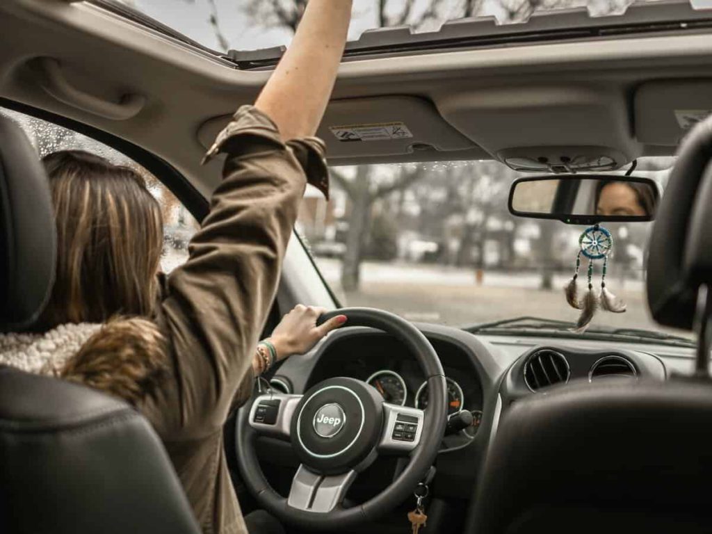 A young female driver has her right arm up through her vehicle’s sunroof as she sits behind the wheel.