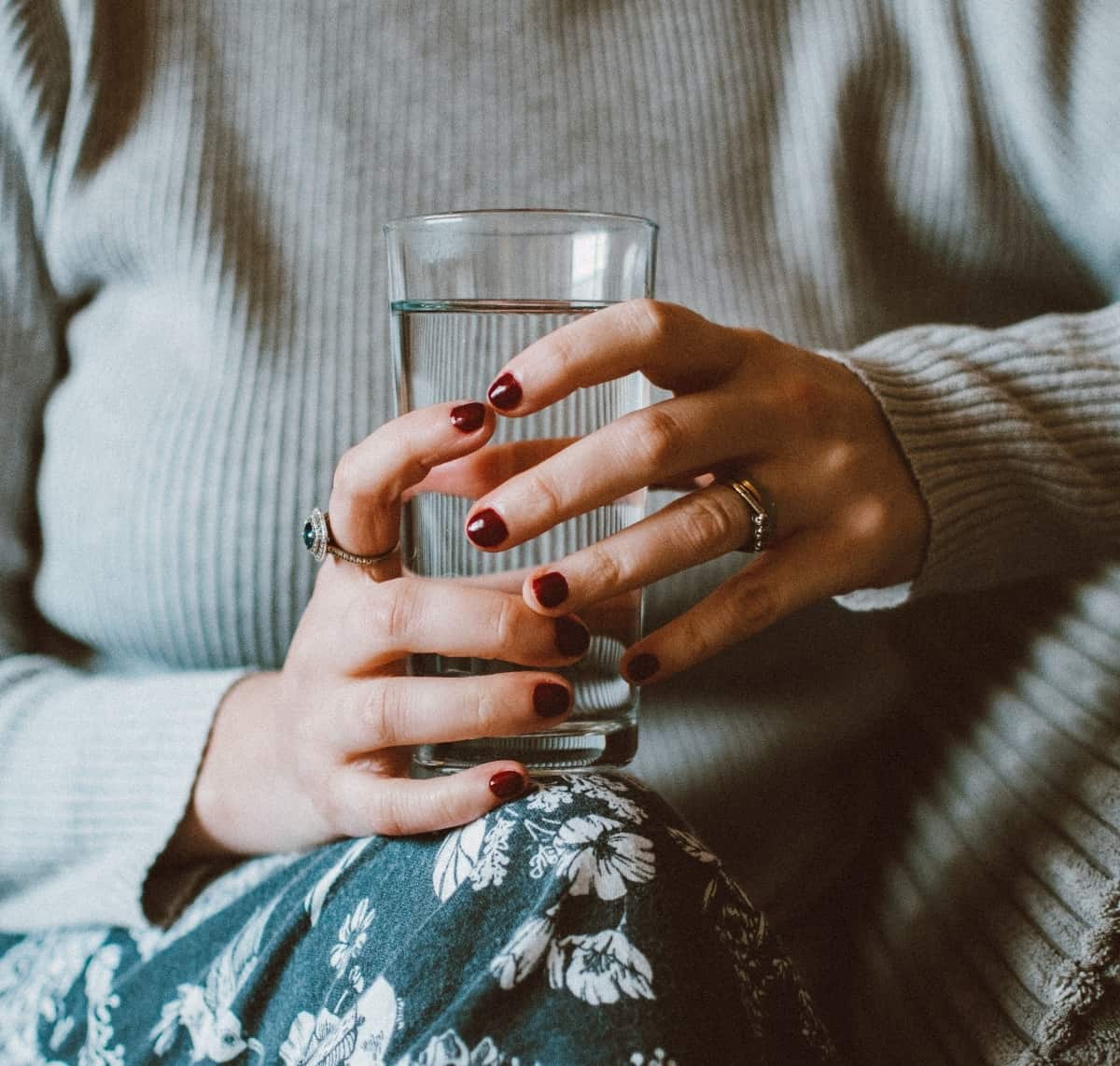 A woman with dark red fingernails is holding a glass of water on her leg.