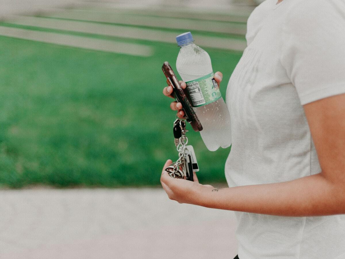 A woman holding a cold water bottle, her cellphone, and dangling car keys walks across a sport's field.