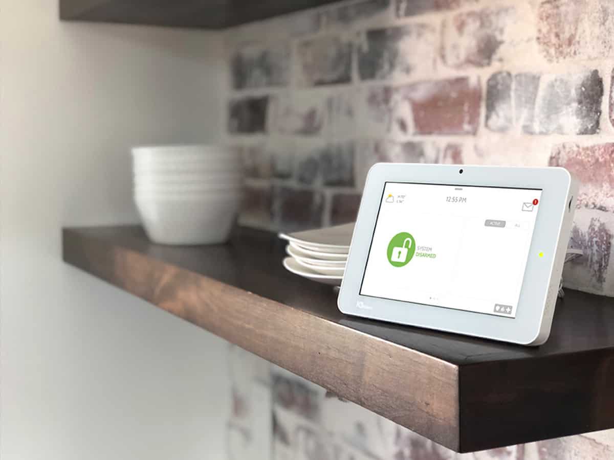 A Qolsys IQ Panel sits on a wooden shelf that's against a brick wall.