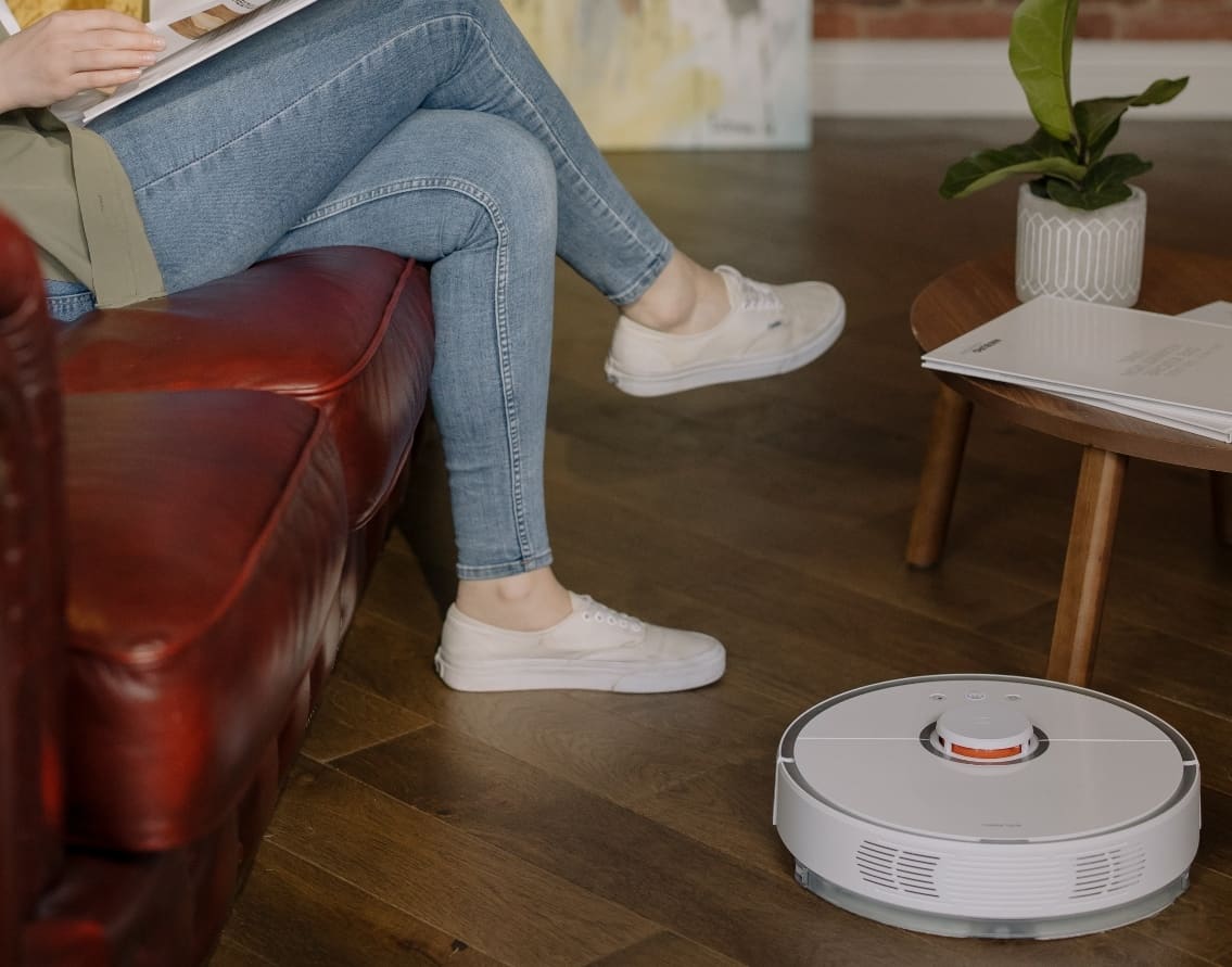 A white robot vacuum sits on the wooden floor next to a woman reading a magazine on a red leather couch.