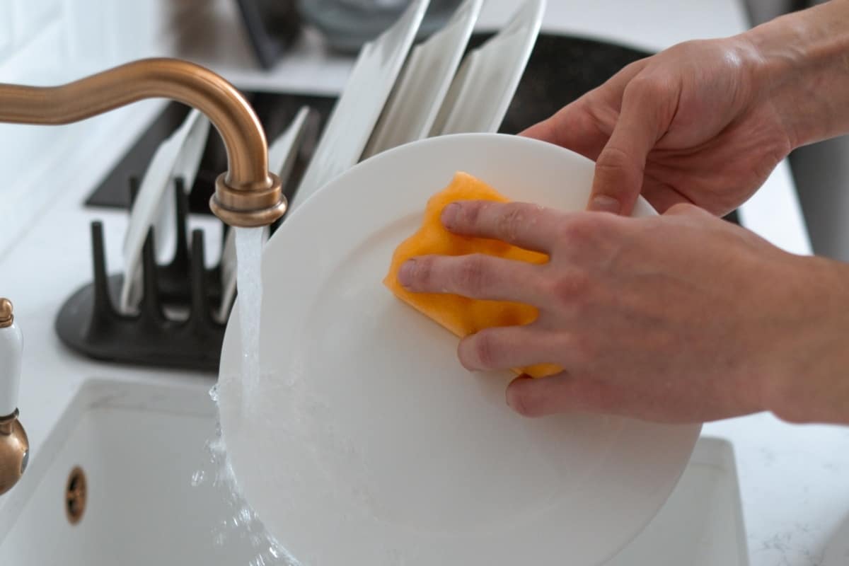 A hand holding a bright orange sponge is wiping a white plate as water flows from a trendy gold faucet.