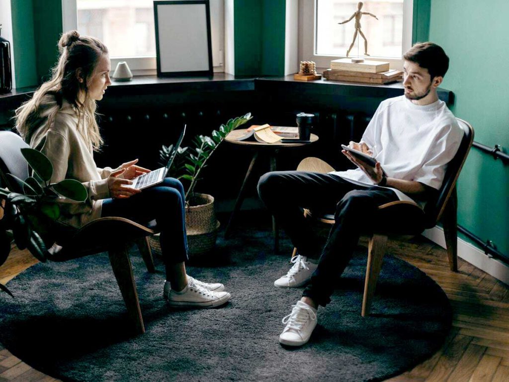 A young couple sits and talks inside their home's living room while holding electronic devices.
