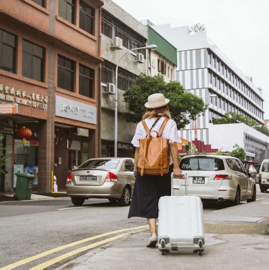 A young woman dressed in stylish clothing walks with a suitcase on the sidewalk in an urban city.