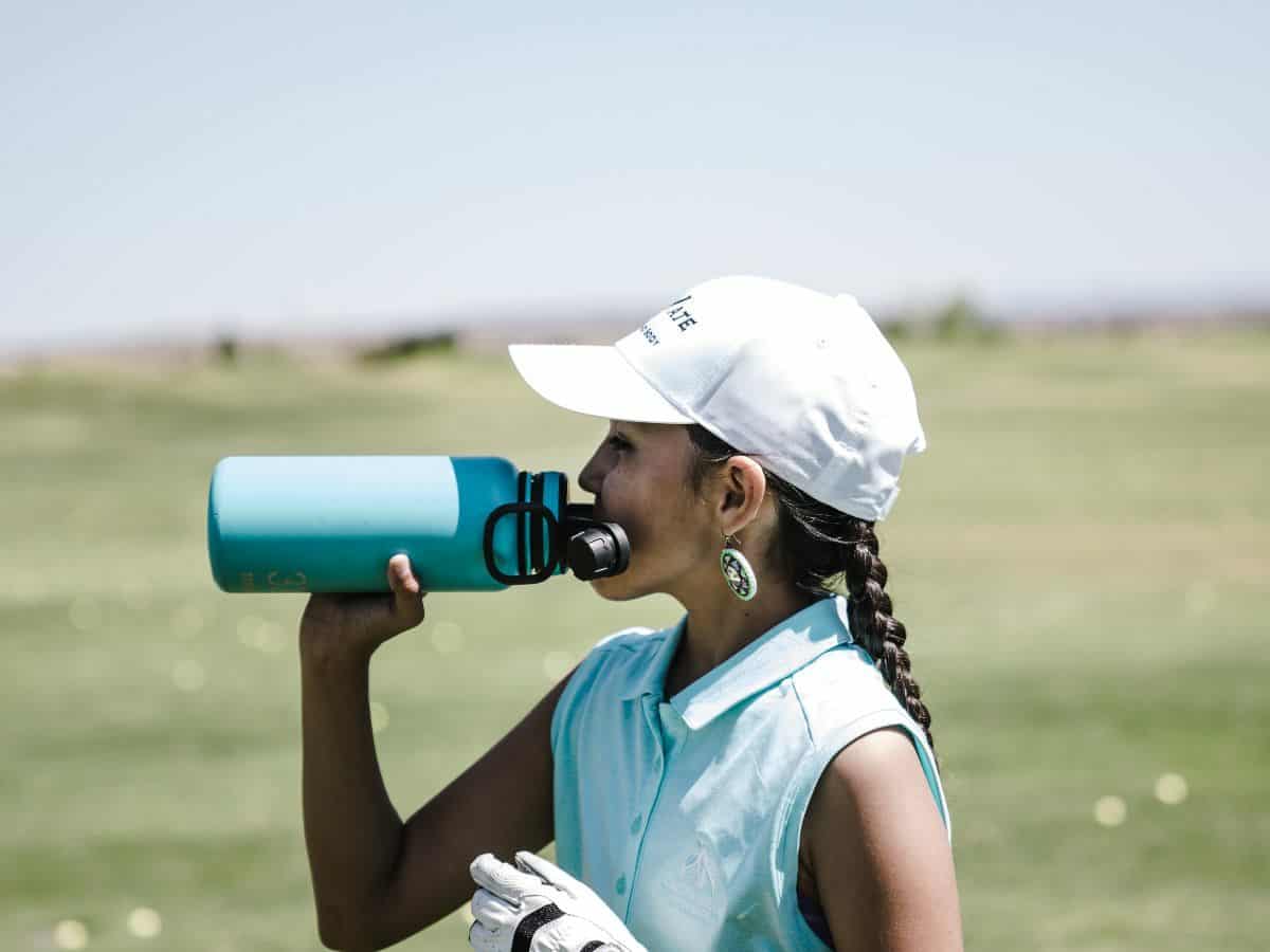 woman golfer in white hat and blue shirt drinking from a blue water bottle