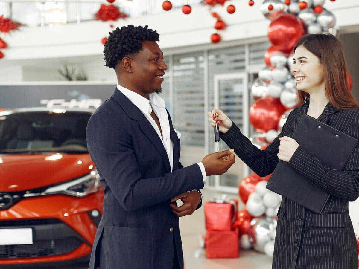 A young man in a suit is being handed car keys from a woman selling the vehicle in the dealership.