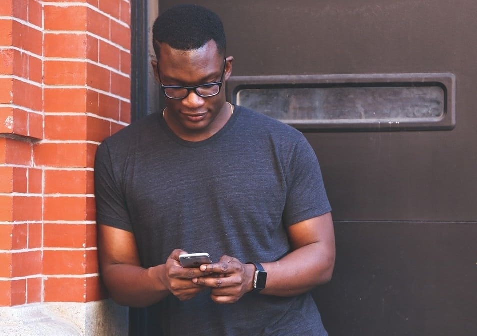 A man wearing glasses looks down at his smartphone as he stands outside next to a red brick building.