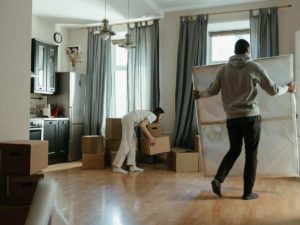 A young woman places a cardboard box onto the wooden floor of a new home while her husband carries in a painting.