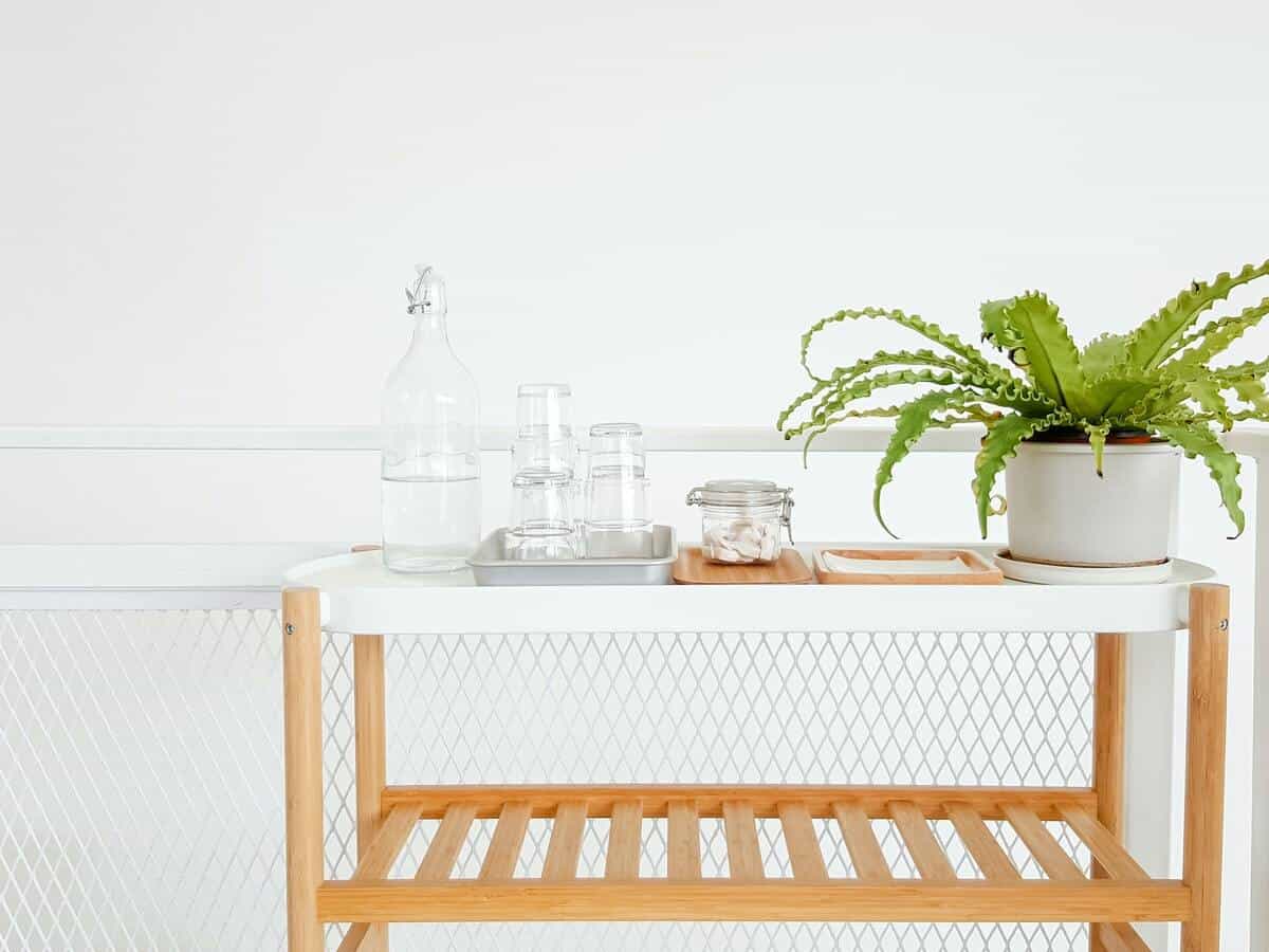 Pitcher of water on a white cart with glasses and a potted house plant.