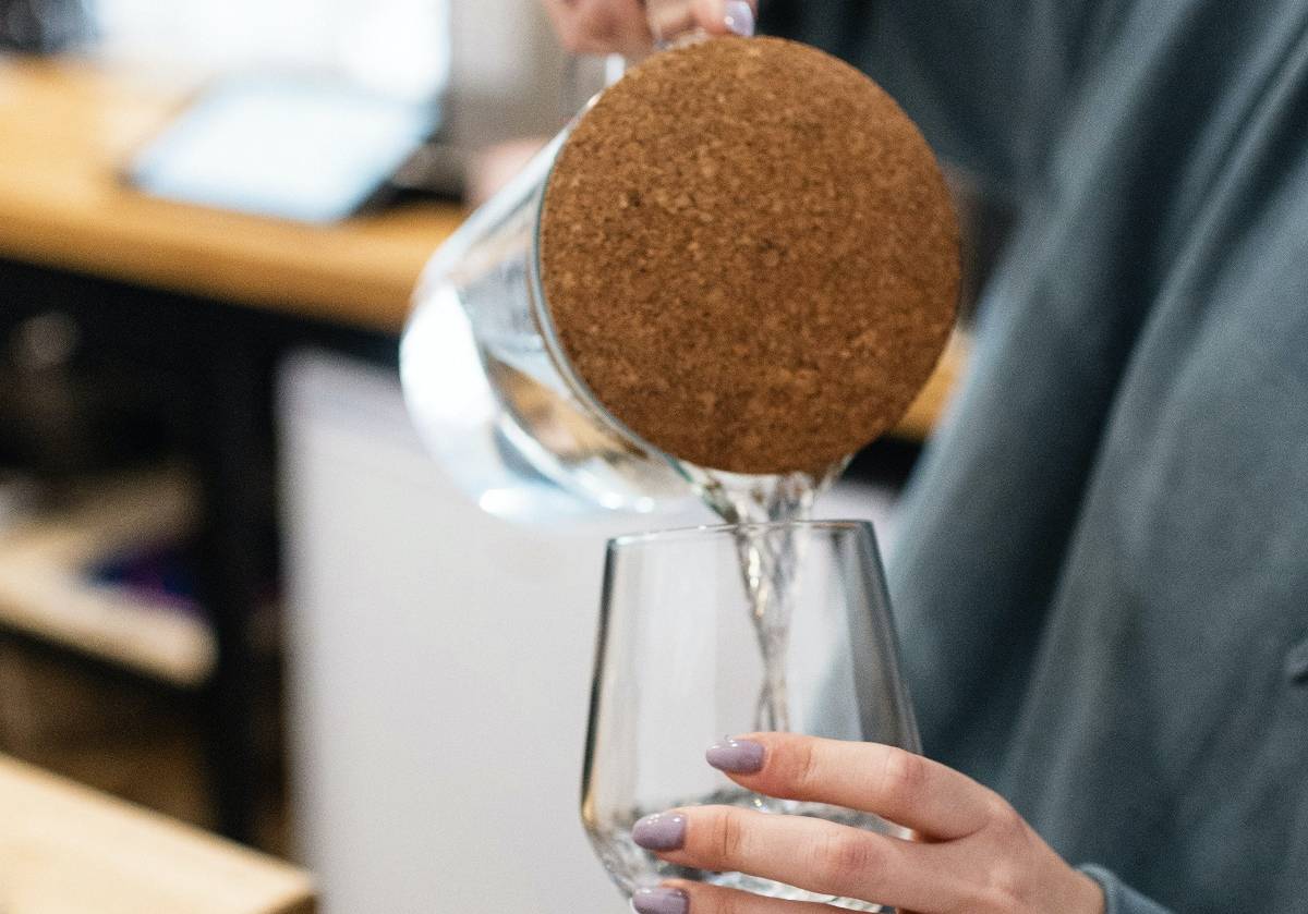 A woman with lavender nail polish on her finger is pouring water into a glass in a kitchen from a glass pitcher with a soft wooden top.