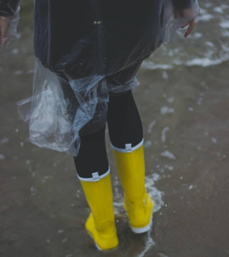 A woman wearing a dark jacket under a clear poncho walks in the rain in bright yellow rain boots.