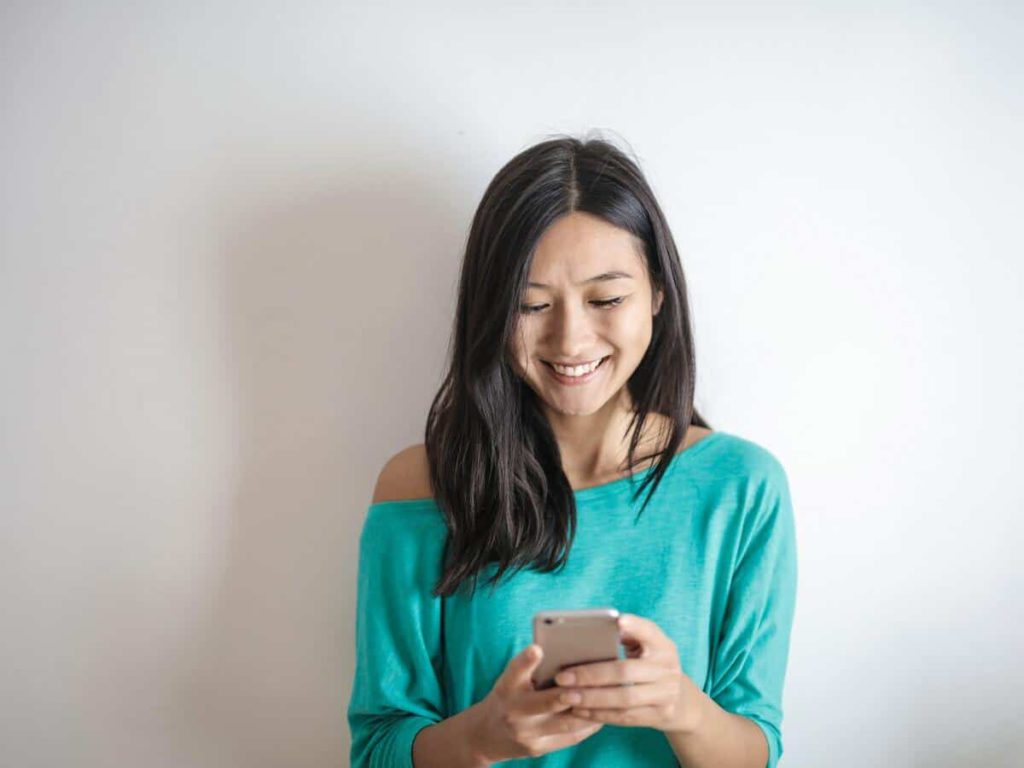 A woman wearing a teal t-shirt is looking down at her smartphone and smiling.
