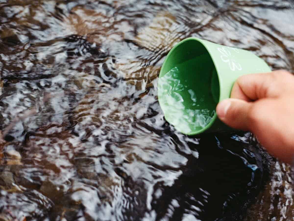 A hand is filling a green cup with water from a spring outdoors.