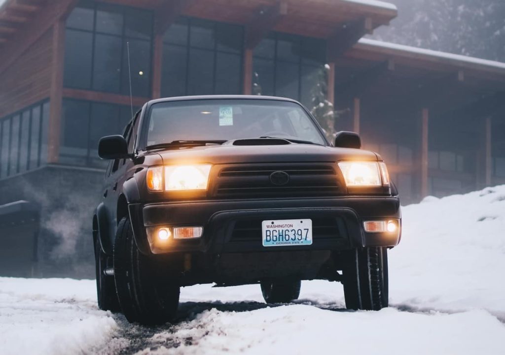 A large SUV with glowing headlights is sitting in snow outside of a rustic home ready to drive off.