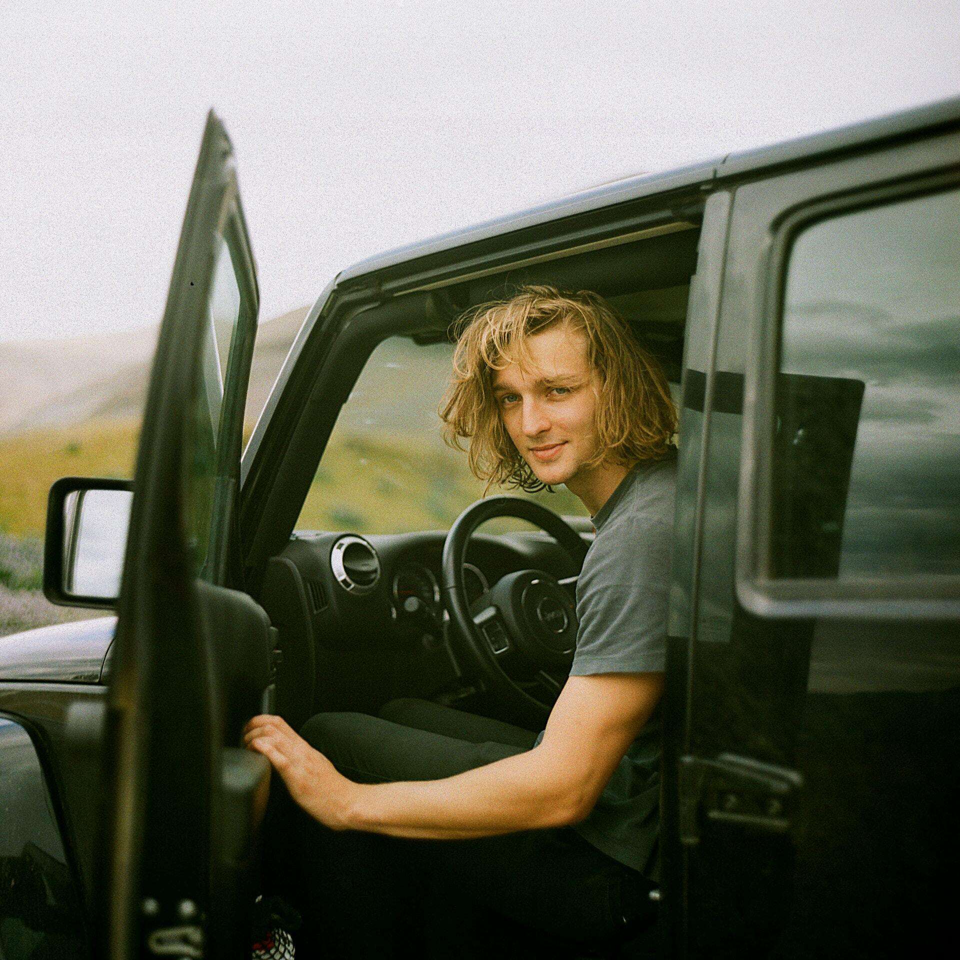 A young man sits with the door open inside of a black used car that’s outdoors.