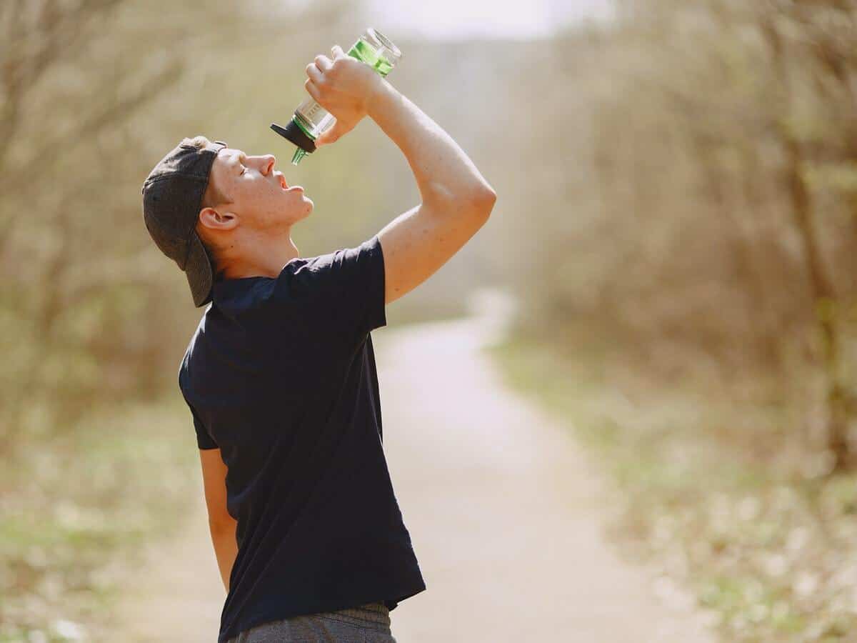 A young man with a baseball cap on backwards drinks from a filtered water bottle in a wooded setting.