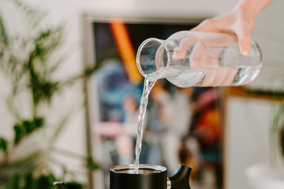 A hand pouring water from a glass carafe into a black teapot kettle.