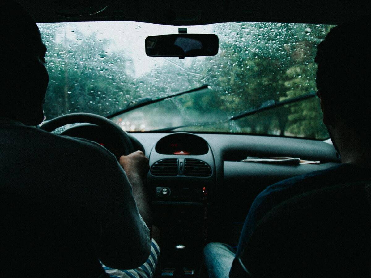Windshield wipers are wiping away rain as two passengers ride inside a vehicle driving down a road.