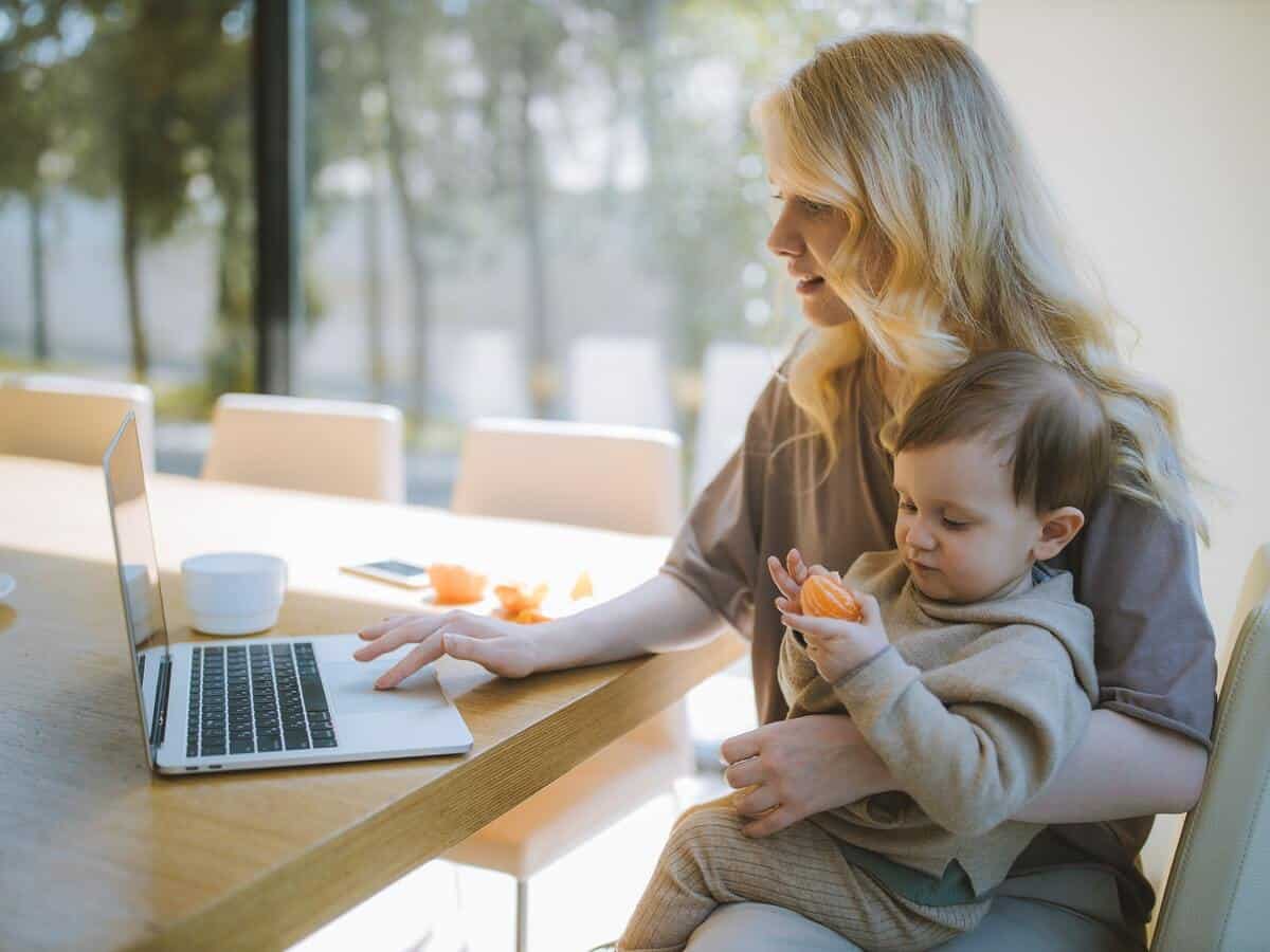 A young woman with blonde hair is holding her baby that’s playing with an orange as she scrolls on her laptop at a kitchen table.