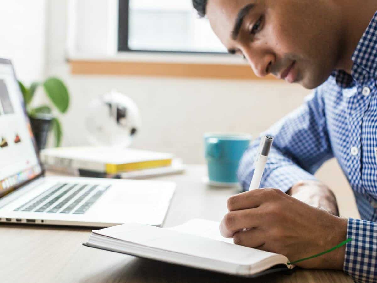 A young man is writing inside of a notebook at a desk holding his laptop, coffee cup, and small stack of books.