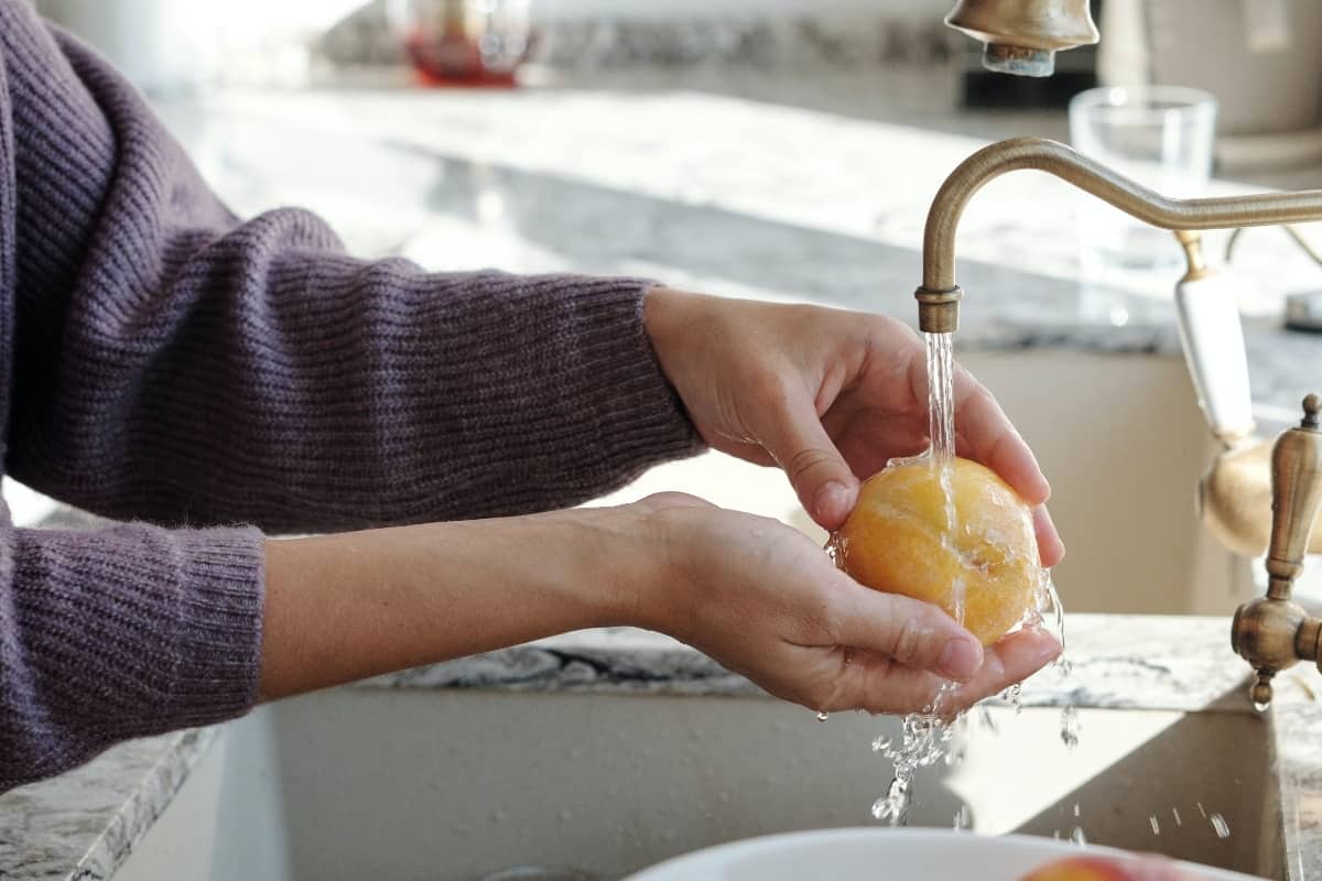 The outstretched arms of a woman rinsing a piece of fruit in a kitchen sink.
