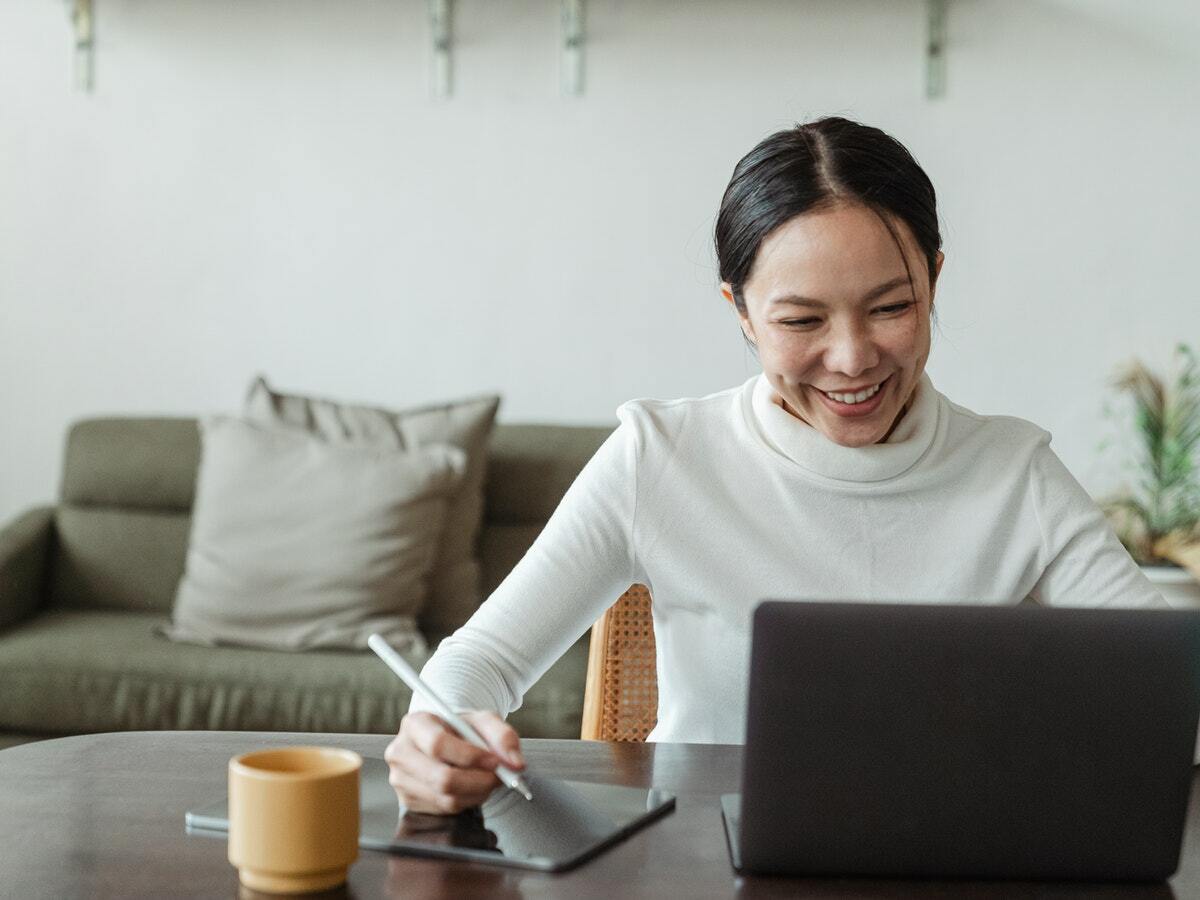 A woman smiles while looking at help laptop and writing on her tablet.