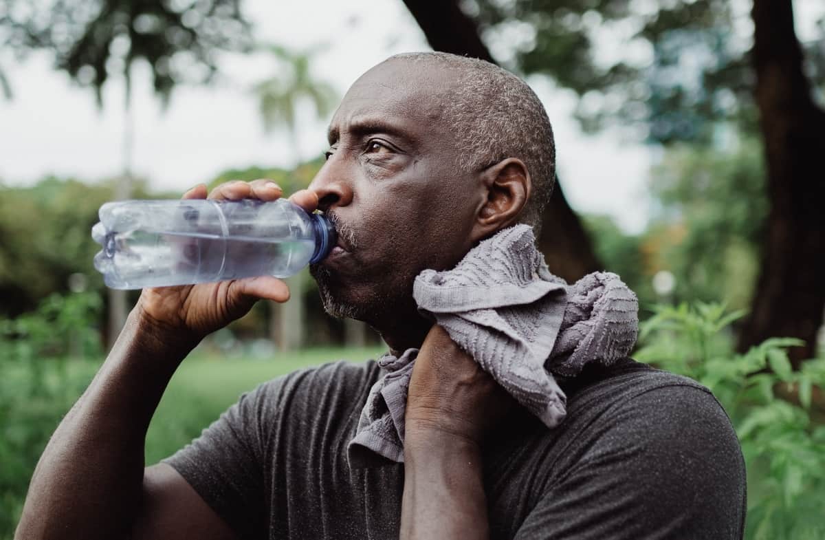 An older man with greying hair is drinking from his water bottle outside as he wipes sweat from off the back of this neck.