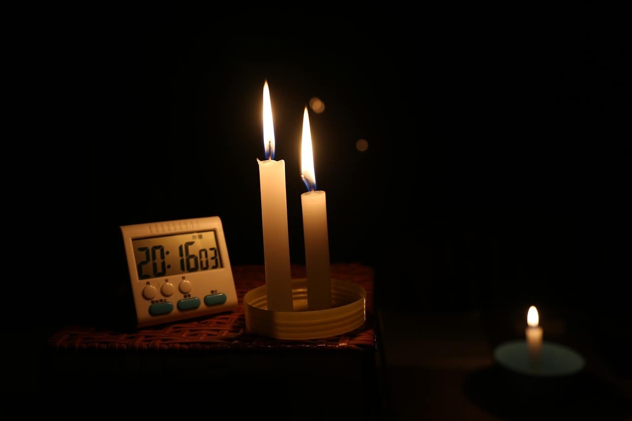 Two lit candles sit on a corner table beside a clock in a dark home.