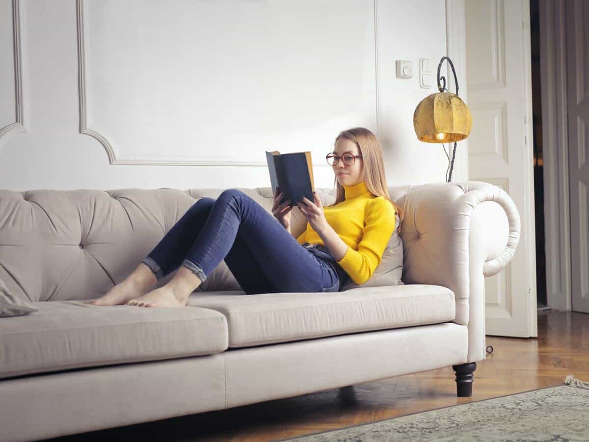 A young woman reading a book on a couch inside of a home.