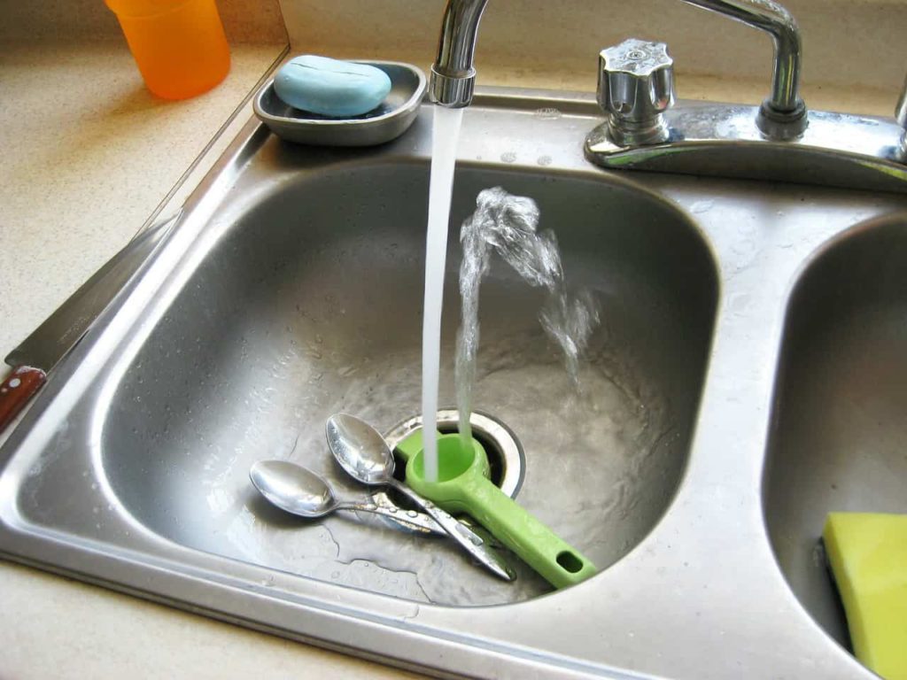 A stainless steel sink with water flowing down from the faucet onto utensils.