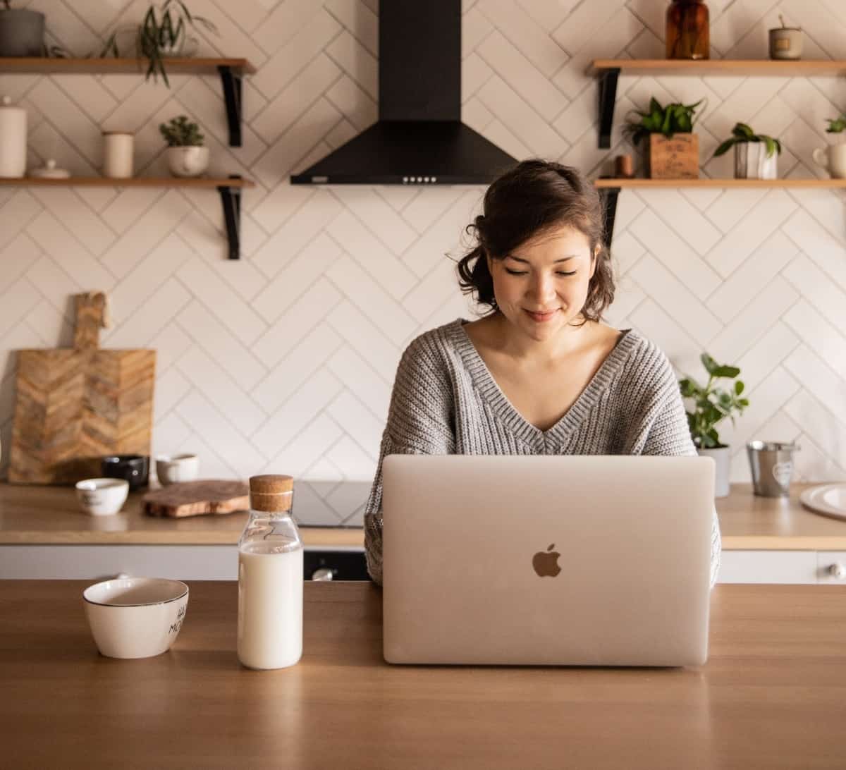 A woman with dark brown hair is smiling as she works on her laptop inside of her kitchen while sitting at a wooden island.