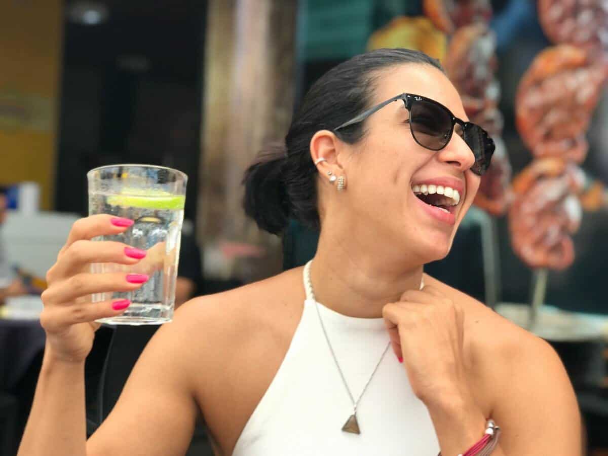 A woman with dark hair is laughing while holding up a glass of water.