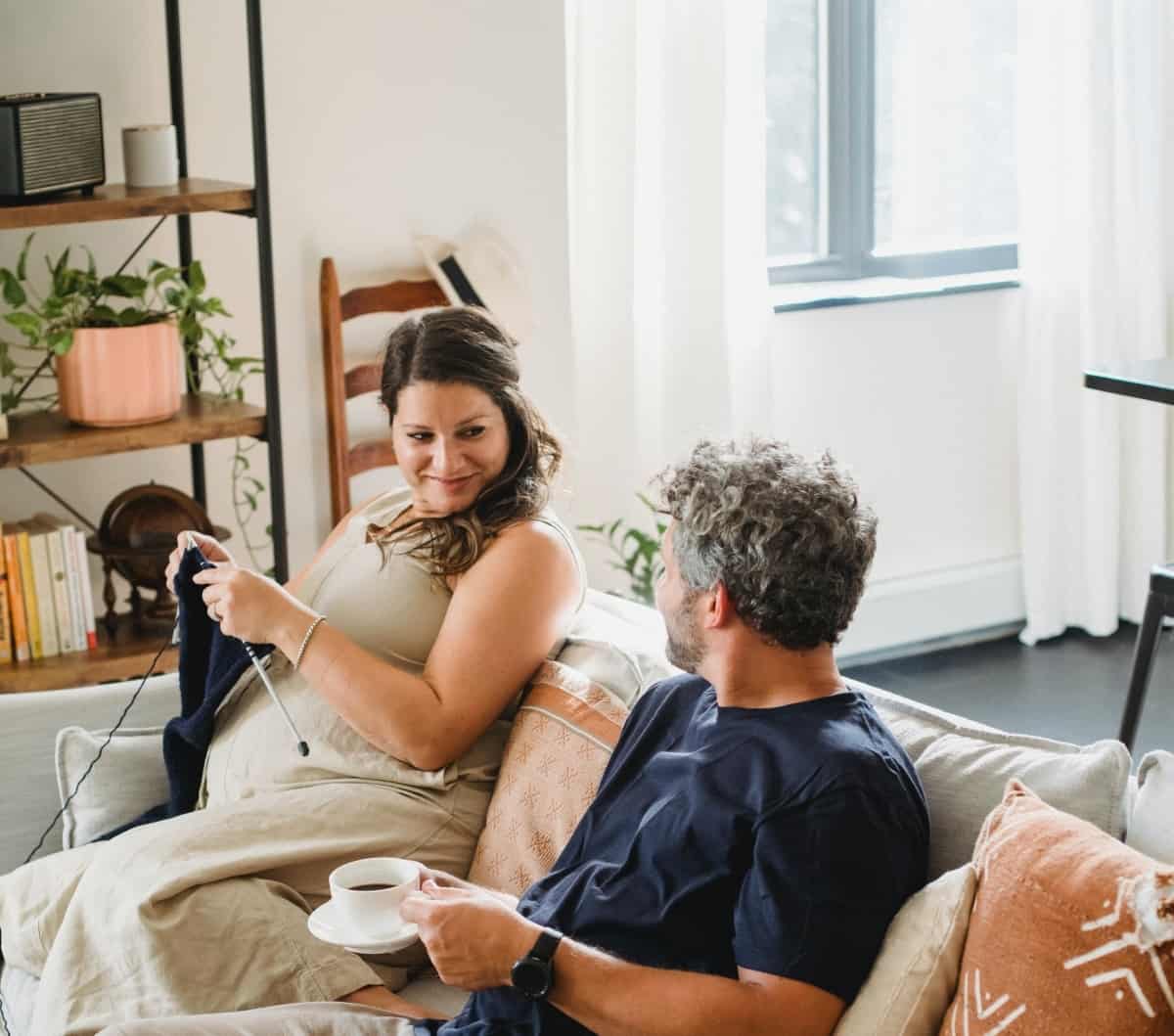 A pregnant woman sits on the couch inside of a living room while knitting next to her husband, who is drinking coffee.