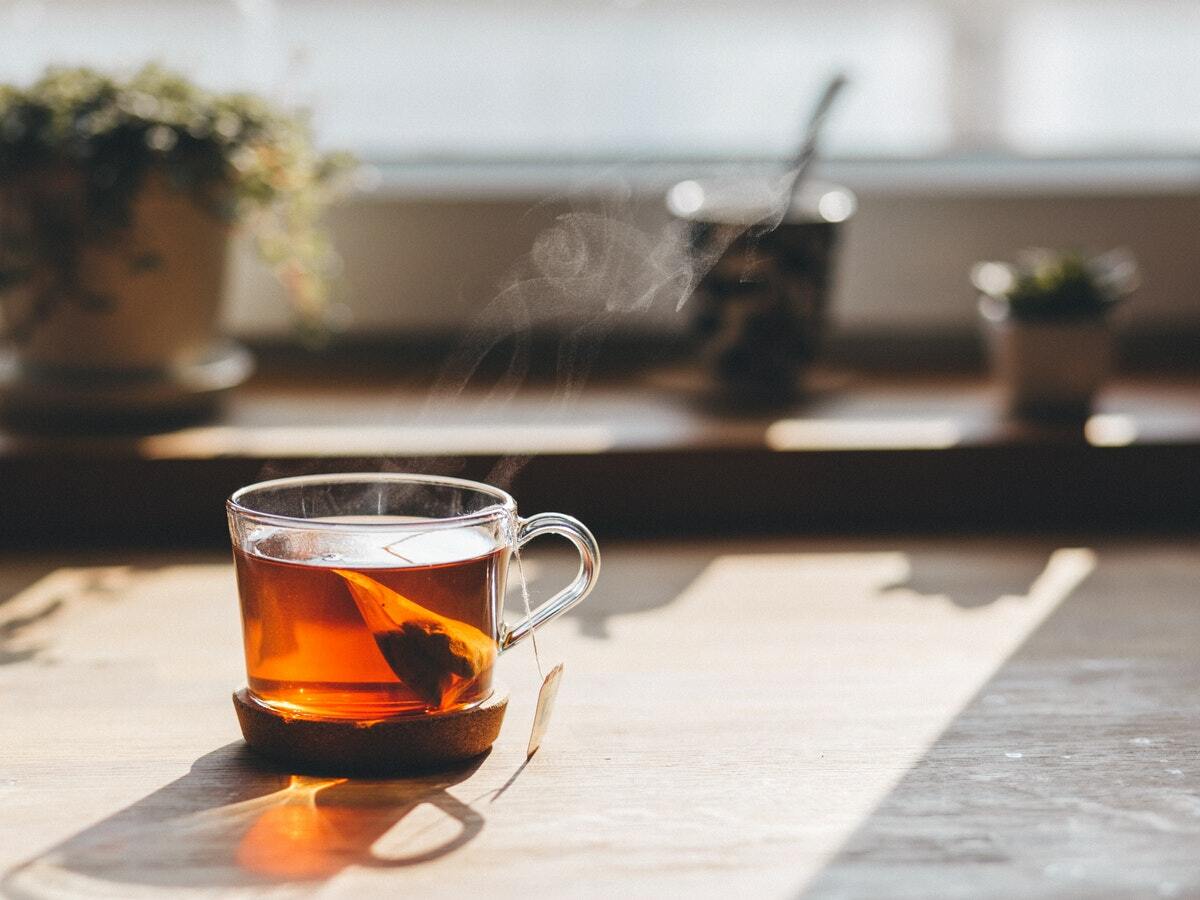 A glass tea cup sitting on a wooden table with a tea bag inside making hot brown tea.