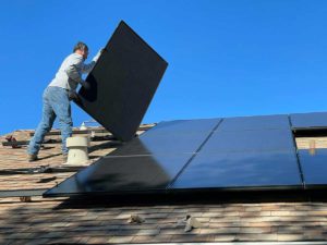 A man is lifting solar panels up over a roof during an installation.