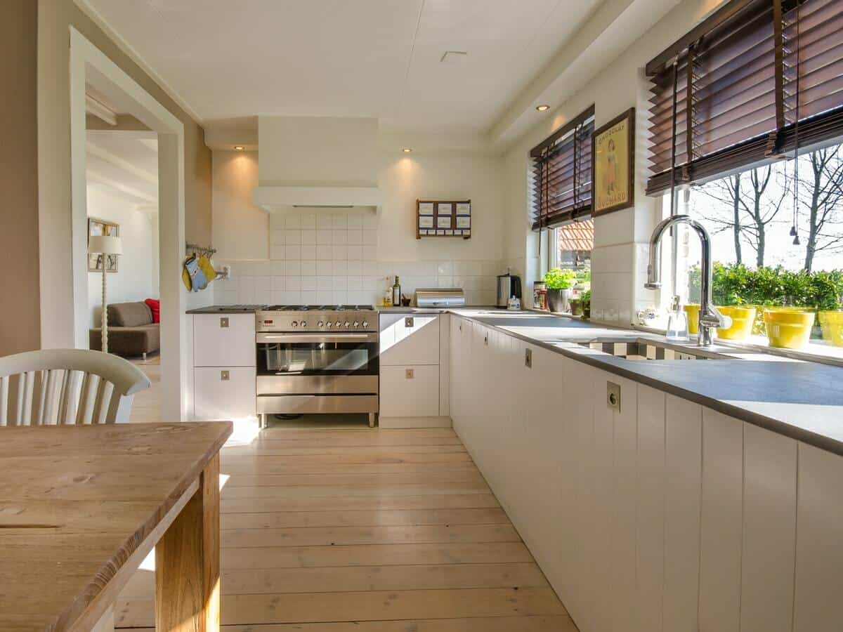 A modern kitchen with white finishes and stainless steel appliances has sunlight glowing inside from the windows.