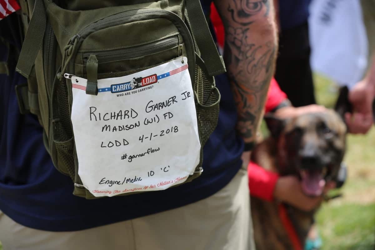 A dark green backpack with a paper attached reading the name of a fallen soldier.