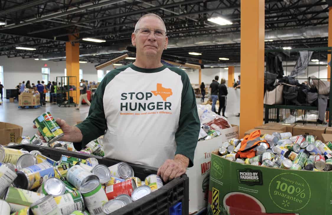 A volunteer at North Texas Food Bank is packing canned food items while wearing a t-shirt that says "Stop Hunger."